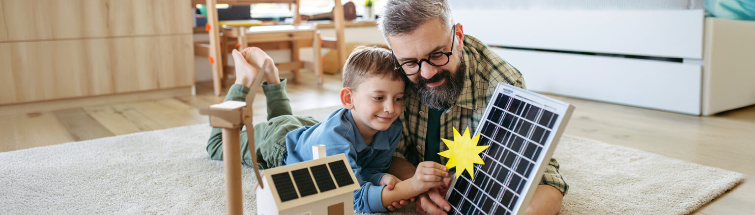 Ein Vater und sein Sohn liegen auf dem Teppich und betrachten spielerisch ein Solarmodell mit Sonnensymbol – Symbolbild für den bewussten Umgang mit Ökostrom und erneuerbarer Energie im Alltag