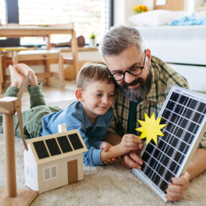 Ein Vater und sein Sohn liegen auf dem Teppich und betrachten spielerisch ein Solarmodell mit Sonnensymbol – Symbolbild für den bewussten Umgang mit Ökostrom und erneuerbarer Energie im Alltag