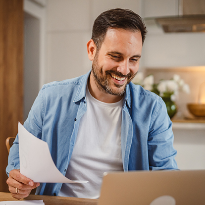 Ein Mann mit Bart sitzt lächelnd am Küchentisch, schaut auf seinen Laptop und hält ein Dokument in der Hand – Symbolbild für den digitalen und flexiblen Stromtarif