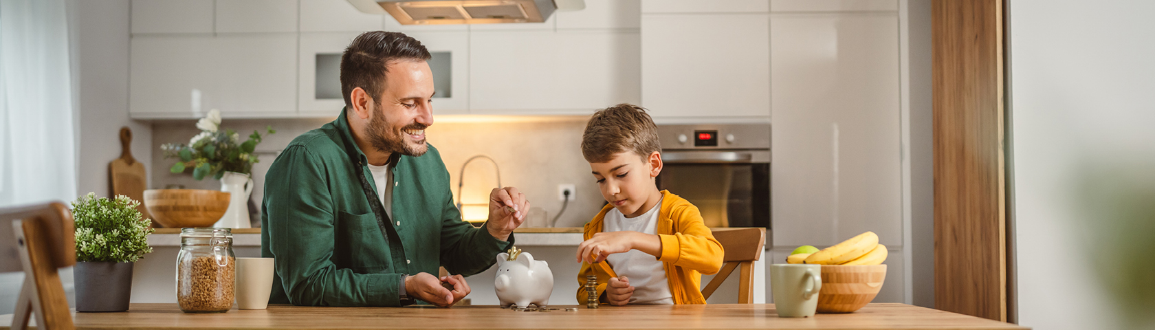 Ein Vater und sein Sohn sitzen am Küchentisch und werfen gemeinsam Geldmünzen in ein Sparschwein – Symbolbild für bewusstes Energiesparen im Alltag