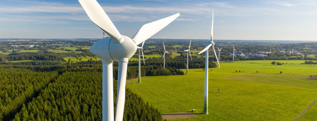 Windkraftanlagen stehen auf grüner Wiese mit Wald und Ort im Hintergrund