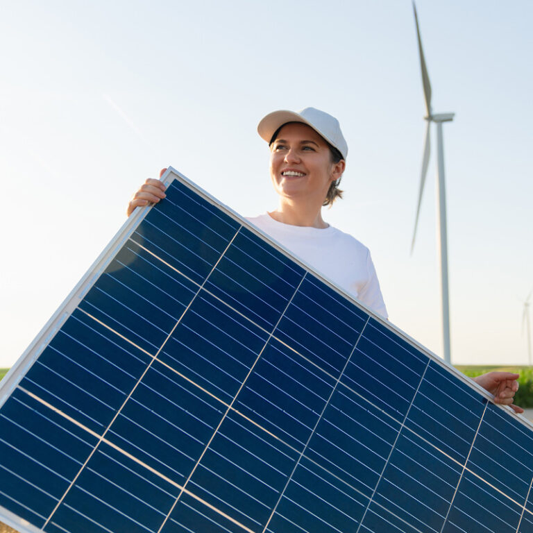 Person hält Photovoltaik-Modul, im Hintergrund Windräder auf offenem Feld