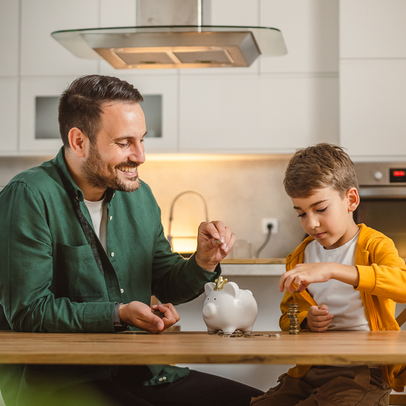 Ein Vater und sein Sohn sitzen am Küchentisch und werfen gemeinsam Geldmünzen in ein Sparschwein – Symbolbild für bewusstes Energiesparen im Alltag